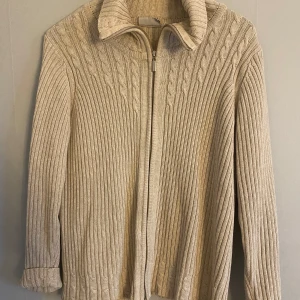 Beige stickad kofta från Cappuccini - Säljer en stilren beige stickad kofta från Cappuccini. Koftan har en dragkedja framtill och ett diskret flätmönster längs med sidorna. Perfekt för kyligare dagar med sin långa ärmar och mjuka material.