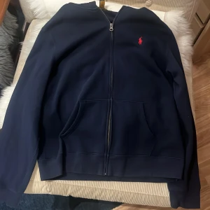 Mörkblå hoodie från Polo Ralph Lauren - Säljer en snygg mörkblå hoodie från Polo Ralph Lauren med dragkedja och broderad röd logga på bröstet. Den har långa ärmar och en praktisk ficka framtill. Perfekt för en avslappnad stil. Använd 2 gånger gott som ny inga defekter på användning storlek 18-20 år sitter som s 