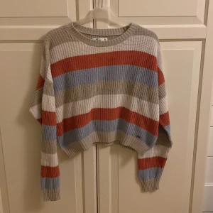 Randig stickad tröja från Hollister - Säljer en mysig stickad tröja med breda ränder i beige, blått och rött. Tröjan har en loose passform och långa ärmar, perfekt för kyliga dagar. Den är ribbad och har en rund halsringning. Kort modell och storleken är M. Från Hollister. I fint skick, men tecken på användning. 