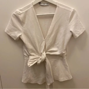 Vit v-ringad topp från MNG Suit - Säljer en stilren vit v-ringad topp från MNG Suit. Toppen har korta ärmar och ett knytband som man knyter runt midjan. INTE MINA BILDER