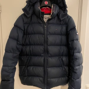 Mörkblå dunjacka från Tommy Hilfiger - Säljer en mörkblå dunjacka från Tommy Hilfiger med huva och dragkedja. Jackan har en klassisk pufferdesign och långa ärmar. Perfekt för kyliga dagar.ett hål på baksidan och vid den högra ärmen därav priset