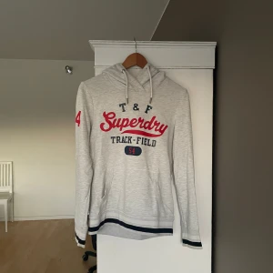 Grå hoodie från Superdry - Säljer en grå hoodie från Superdry med röd och marinblå text på framsidan. Den har en justerbar huva med snören och en stor ficka framtill.  Den är även i väldigt bra skick. Nypris är ca 500 kr men säljer den för 200. Priset kan diskuteras vid snabb affär. Hör gärna av dig om du har frågor eller funderingar.