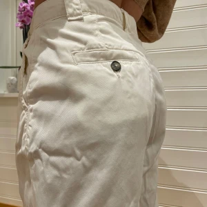 Vita byxor med knappdetalj - Snygga vita byxor med hög midja och en stilren knappdetalj på bakfickan. Byxorna har en rak passform och är perfekta för en avslappnad look. Tillverkade i ett mjukt och bekvämt material.