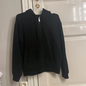 Svart hoodie med dragkedja - Säljer en klassisk svart hoodie med dragkedja och huva. Tröjan har långa ärmar och praktiska fickor framtill. Perfekt för en avslappnad stil.
