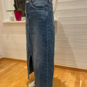 Jeanskjol med slits - Snygg jeanskjol i blå denim med en stilren slits framtill. Kjolen har en klassisk femficksdesign och knappar i midjan. Perfekt för en avslappnad look.