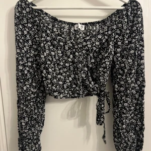 Svart blommig topp från Hollister - Säljer en söt svart blommig topp från Hollister i storlek XS. Toppen har långa ärmar och en knytning framtill. Perfekt för en stilren look med en touch av blommigt mönster.