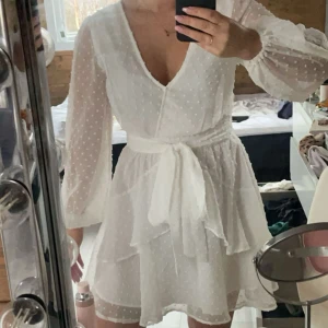 Vit klänning - Vit klänning med volanger och band i midjan. Fint prickigt mönster, genomskinliga ärmar och resterade har dubbelt tyg. Dragkedja i ryggen. Perfekt till student🥰använd 1 gång