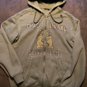 Grön hoodie med text och motiv - Säljer en grön hoodie med dragkedja från Bling Lucid. Den har texte i strass n 'CHASE THE BAG' och är märket Blinglucid. Nypris 600kr. 