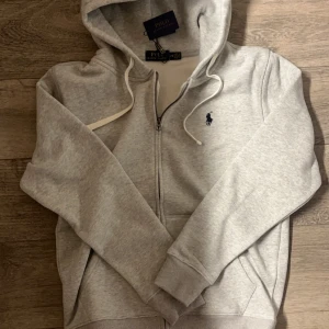  Ralph Lauren zip hoodie  - En modern grå zip-up hoodie i storlek M med mjukt foder som håller dig bekväm och varm under kyliga vinterdagar och friska vårkvällar. Hör av dig om du vill veta mer!