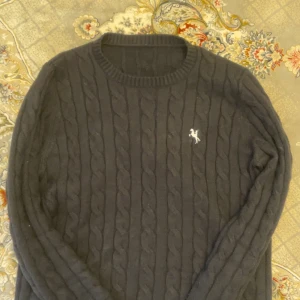 Ralph Lauren Tröja  - Säljer en stilren svart kabelstickad tröja med en liten vit logotyp på bröstet. Tröjan har långa ärmar och en rund halsringning. Perfekt för kyligare dagar och ger en klassisk look.