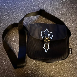 Svart axelväska  - Snygg svart axelväska med ett coolt spänne framtill. Väskan har en justerbar axelrem med text och en praktisk storlek för vardagsbruk. Perfekt för att bära dina nödvändigheter med stil.