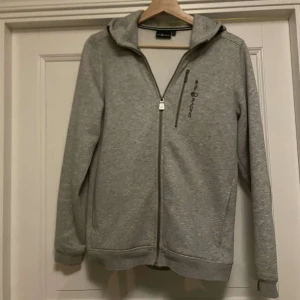 Grå huvtröja från Sail Racing - Säljer en grå huvtröja från Sail Racing med dragkedja och en bröstficka. Tröjan har en broderad logga på bröstet och huvan. Tillverkad i en mjuk bomulls- och polyesterblandning. Perfekt för en avslappnad stil.