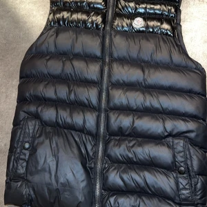 Svart dunväst från Moncler - Snygg svart dunväst från Moncler med dragkedja och två fickor framtill. Insidan är i en kontrasterande gul färg. Perfekt för kyligare dagar när du vill hålla stilen. Pris kan diskuteras 