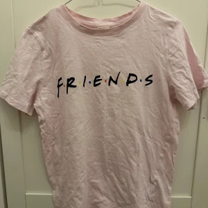 Rosa t-shirt med Friends-tryck - Säljer en rosa t-shirt med det ikoniska Friends-trycket på framsidan. Perfekt för alla fans av den klassiska TV-serien. T-shirten är kortärmad och gjord i mjukt material för en bekväm passform.❤️💛💙