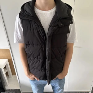 Svart dunväst med huva - Snygg svart Zara dunväst med huva och dragkedja. Västen har knappar framtill och praktiska fickor.