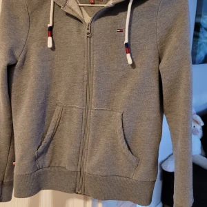Grå hoodie från Tommy Hilfiger - Säljer en snygg och bekväm grå hoodie från Tommy Hilfiger Denim i storlek XXS. Den har en dragkedja framtill och praktiska fickor. Perfekt för en avslappnad stil och passar bra till både jeans och leggings. Huvan har justerbara snören med färgdetaljer. 🩶
