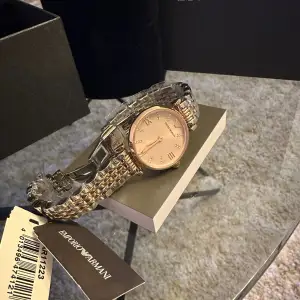 Säljer en elegant guldfärgad klocka från Emporio Armani. Klockan har en stilren design med en glittrig urtavla och romerska siffror. Armbandet är i metall och ger en lyxig känsla. Perfekt för både vardag och fest!