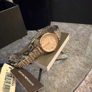 Guldfärgad klocka från Emporio Armani - Säljer en elegant guldfärgad klocka från Emporio Armani. Klockan har en stilren design med en glittrig urtavla och romerska siffror. Armbandet är i metall och ger en lyxig känsla. Perfekt för både vardag och fest!