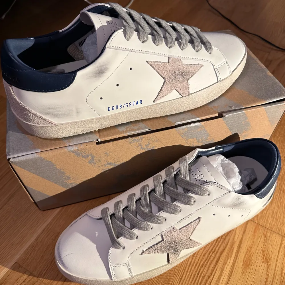 Säljer ett par snygga Golden Goose sneakers i vitt med grå stjärndetalj på sidan. Skorna har en cool vintage-look med grå snörning och mörkblå häl. Perfekta för både vardag och fest! 🕺. Skor.