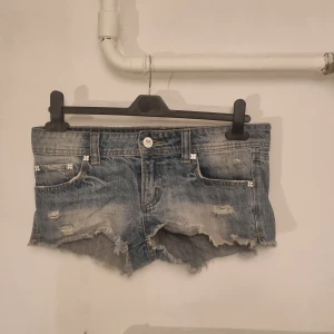 Jeansshorts med slitningar - Säljer ett par jeansshorts i blått med slitningar och fransiga kanter. De har en låg midja och är dekorerade med små blommor vid fickorna. 
