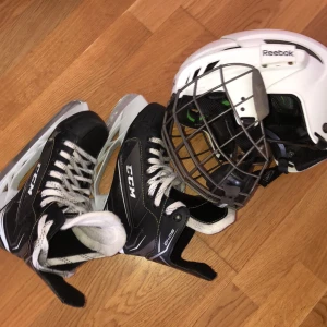 Hockeyutrustning med hjälm och skridskor - Säljer en vit Reebok hockeyhjälm med galler och ett par svarta CCM skridskor. Hjälmen har justerbar passform och skridskorna är i bra skick med snörning. Perfekt för vinterns hockeymatcher eller träning på isen! 🏒