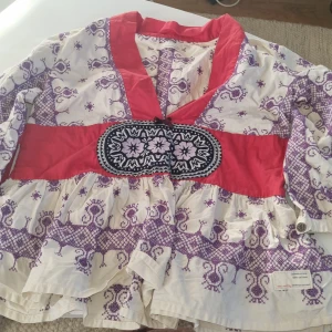 Mönstrad blus från Odd Molly - Säljer en unik blus från Odd Molly i mycket bra skick. Den har ett vackert lila mönster på vit bakgrund med röda detaljer och ett brett midjeband. Klänningen har volanger och en knytning i midjan för en fin passform. Perfekt för vår och sommar!