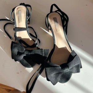 Svarta klackskor med rosett - Snygga svarta klackskor med en stor rosett framtill och smala remmar som knyts runt ankeln. Perfekta för sommarens fester eller en elegant kväll ute. De har en bekväm klack och är i mycket bra skick. Vet inte storleken exakt men skulle gissa på 36/37