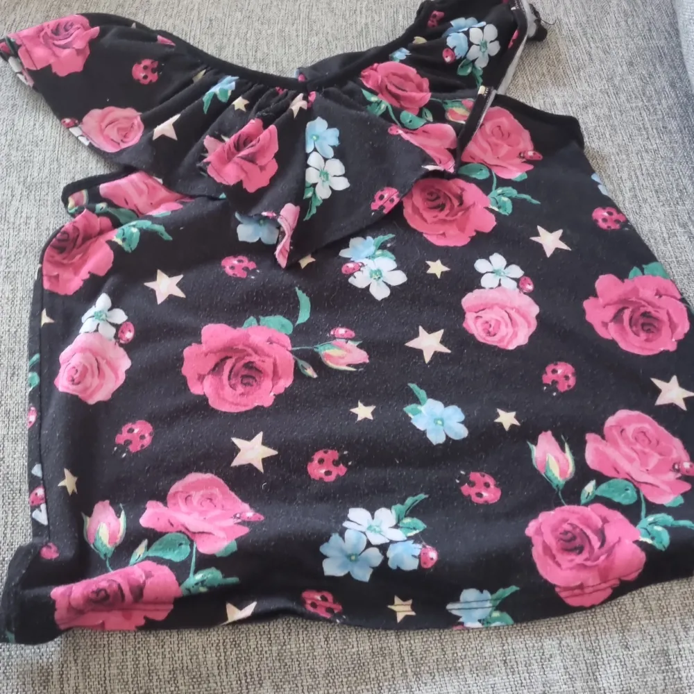 Säljer en superfin offshoulder topp från Justice med ett färgglatt blommönster. Toppen är svart med stora rosa rosor, små vita blommor och stjärnor. Den har en volangdetalj vid halsen som ger en söt touch. Perfekt för sommarens fester eller en dag på stan! 🌸. Toppar.