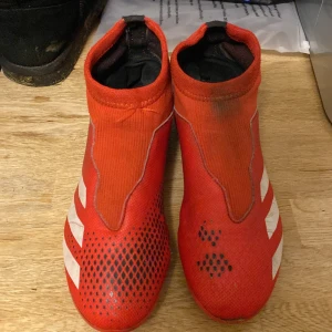 Röda Adidas Predator fotbollsskor - Säljer ett par röda Adidas Predator fotbollsskor i bra skick. De har en snygg design med vita detaljer och är perfekta för fotbollsträning. Skorna har en tight passform och är gjorda i syntetmaterial för bästa prestanda på planen.