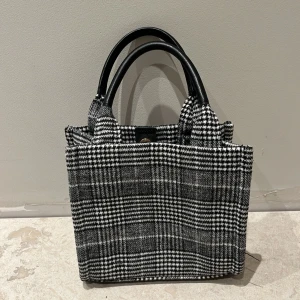 Rutig handväska med svarta handtag - Snygg grå och vit rutig handväska från H&M. Väskan har en klassisk design med ett rymligt inre och en praktisk knappstängning. Den är i mycket bra skick och redo att användas direkt!