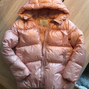 Rosa dunjacka från Woolrich - Säljer en superfin glansig rosa pufferjacka från Woolrich i storlek 12 år. Den har en stor huva, dragkedja framtill samt två fickor med dragkedja.  Små små skiftningar i glansen kan ses i visst ljus men inget som stör (märks knappt) samt ett ytligt skrapmärke på ryggen, knappt synbart (se sista bilden)  i övrigt mycket gott skick. Inköpt 2023, nypris 4100kr