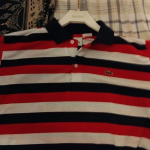 Randig pikétröja från Lacoste - Säljer en snygg randig pikétröja från Lacoste i rött, vitt och blått. Tröjan har en klassisk krage och knappar framtill. Perfekt för en avslappnad men stilren look. Passar både till vardags och festliga tillfällen.