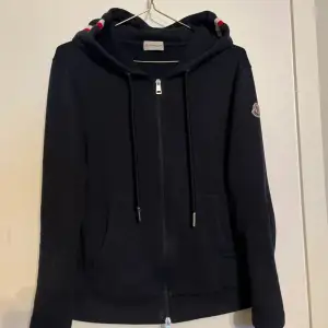 Säljer en snygg svart hoodie från Moncler i mycket bra skick. Den har en dragkedja framtill och en klassisk Moncler-logga på ärmen. Perfekt för höst och vår, med en skön passform och praktiska fickor. Huvan har en stilren design med snören. och är nästan helt ny och äkta Hör av er vid funderningar.