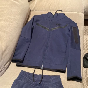 Nike tech fleece tracksuit - Säljer ett mörkblått träningsset från Nike i mycket bra skick. Setet består av en hoodie med dragkedja och matchande byxor. Perfekt för träning eller vardagsbruk. Materialet är mjukt och bekvämt, med elastisk midja på byxorna för bästa passform.
