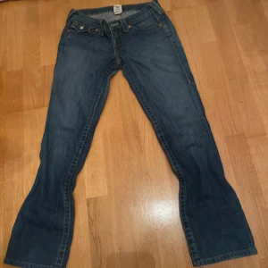 Blå jeans från True Religion - Säljer ett par snygga blå jeans från True Religion i storlek 27. De har en klassisk femficksdesign med coola detaljer på bakfickorna. Jeansen är i bra skick och perfekta för både vardag och fest. Passformen är normal och de är tillverkade i USA. Perfekt för dig som vill ha en stilren look! 👖