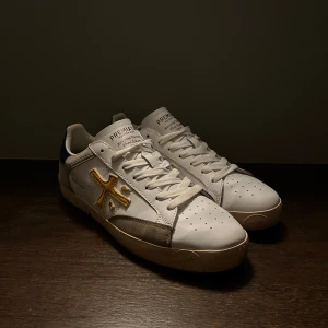 Vita sneakers från Premiata - Säljer ett par vita Premiata sneakers i bra skick. De har en snygg gul detalj på sidan och blå detaljer på hälen. Skorna har en klassisk design med snörning och är perfekta för vardagsbruk. Sulan är i gott skick och de är bekväma att gå i. Perfekta för dig som vill ha stil och komfort i ett!