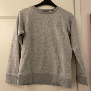 Grå sweatshirt från GANT - Säljer en stilren grå sweatshirt från GANT i mycket bra skick. Tröjan har en klassisk rund halsringning och ett diskret GANT-emblem på bröstet. Perfekt för både vardag och skola. Superbekväm och lätt att matcha med andra plagg!