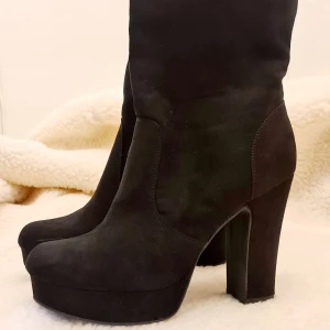 Svarta mockastövletter med klack - Snygga svarta stövletter i mocka med hög klack och platåsula. Perfekta för höst och vinter, dessa boots ger en elegant touch till vilken outfit som helst. De är i bra skick och har en robust klack som mäter cirka 9 cm. Passar både till vardags och festliga tillfällen.