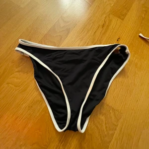 Svart bikiniunderdel från Intersport - Säljer en snygg svart bikiniunderdel från Intersport i storlek 36. Den har vita kontrastkanter som ger en stilren look. Materialet är 84% polyamid och 16% elastan, vilket gör den både bekväm och stretchig. Perfekt för sommarens alla badäventyr! 🖤