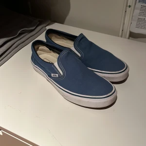 Blå slip-on sneakers från Vans - Säljer ett par klassiska blå slip-on sneakers från Vans. De har en vit sula och är i bra skick med lite slitage på sulan. Perfekta för en avslappnad stil och passar till det mesta. Skick 7.5/10