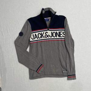 Säljer en snygg grå och marinblå tröja från Jack & Jones. Den har en dragkedja vid halsen och ett stort tryck med märkesnamnet över bröstet. Tröjan har långa ärmar och ribbade muddar med röd och marinblå rand. Perfekt för både fina och avslappnade tillfällen. Strl S