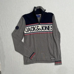 Grå och marinblå tröja från Jack & Jones - Säljer en snygg grå och marinblå tröja från Jack & Jones. Den har en dragkedja vid halsen och ett stort tryck med märkesnamnet över bröstet. Tröjan har långa ärmar och ribbade muddar med röd och marinblå rand. Perfekt för både fina och avslappnade tillfällen. Strl S