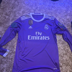 Lila långärmad fotbollströja från Real Madrid (ronaldo) - Säljer en snygg lila långärmad fotbollströja från Adidas med 'Fly Emirates' tryck och Real Madrid-emblem. Tröjan har vita detaljer och nummer 7 med 'Ronaldo' på ryggen. Perfekt för fans! 🌟
