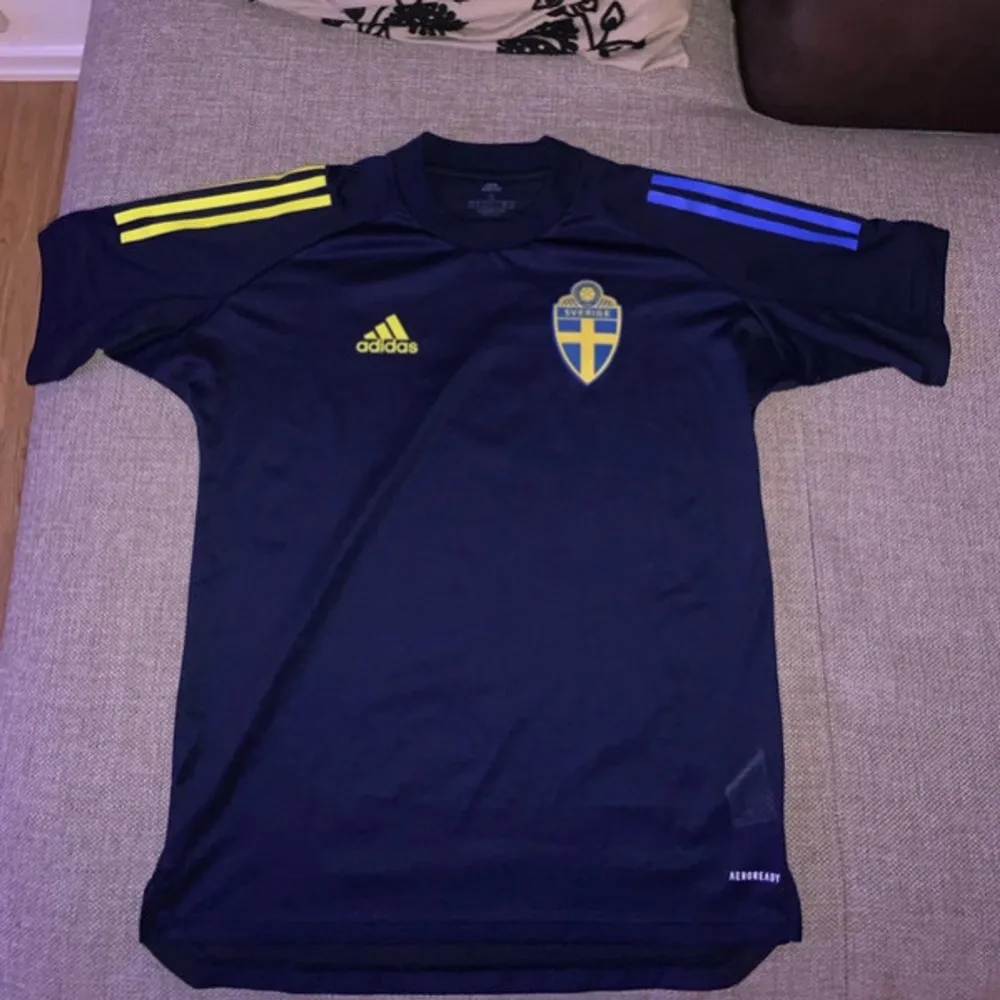 Säljer en snygg svensk landslagströja från Adidas i mörkblått med gula och blå detaljer. Tröjan har korta ärmar och är i AEROREADY-material för bra andningsförmåga. Perfekt för fotbollsfans som vill visa sitt stöd för Sverige!. T-shirts.