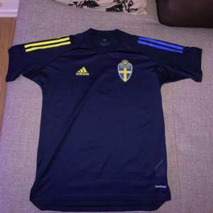 Säljer en snygg svensk landslagströja från Adidas i mörkblått med gula och blå detaljer. Tröjan har korta ärmar och är i AEROREADY-material för bra andningsförmåga. Perfekt för fotbollsfans som vill visa sitt stöd för Sverige!