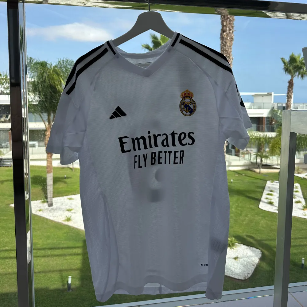 Säljer en vit Real Madrid fotbollströja från Adidas med korta ärmar. Tröjan har svarta detaljer och klubbens emblem på bröstet. På baksidan står det 'MBAPPÉ' och nummer 9. Perfekt för alla fotbollsfans!. T-shirts.