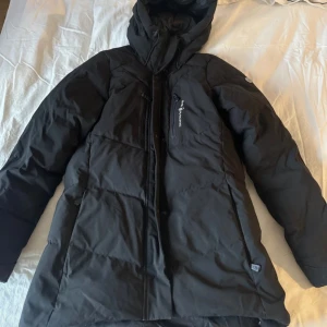 Svart dunjacka från Sail Racing - Säljer en svart dunjacka från Sail Racing i storlek small. Jackan är perfekt för kalla vinterdagar med sin varma och vadderade design. Den har en praktisk huva och flera dragkedjefickor framtill. Jackan är i nyskick och har en stilren look som passar till det mesta. Perfekt för både vardag och utomhusaktiviteter!