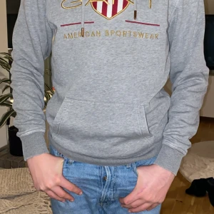 Grå hoodie från GANT - Säljer en snygg grå hoodie från GANT med deras klassiska logga i rött och guld på bröstet. Den har en känguruficka och ribbade muddar vid ärmslut och nederkant. Perfekt för en avslappnad stil!