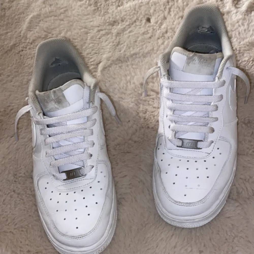 Säljer ett par klassiska vita Nike Air Force sneakers. De är i skinn och har den ikoniska Nike-loggan på sidan. Skorna har snörning och en bekväm passform, perfekt för vardagsbruk. Lite slitna men fortfarande i bra skick.. Kengät.