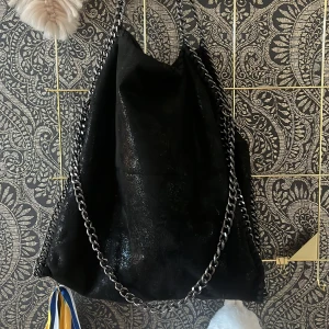Svart väska med kedjedetaljer - Säljer en snygg svart väska med coola kedjedetaljer. Den har en glittrig yta som ger en extra touch av glamour. Perfekt för både vardag och fest!